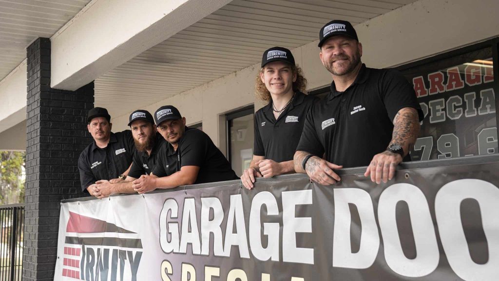 Five Trinity technicians in branded caps and polo shirts standing behind a Trinity garage door banner